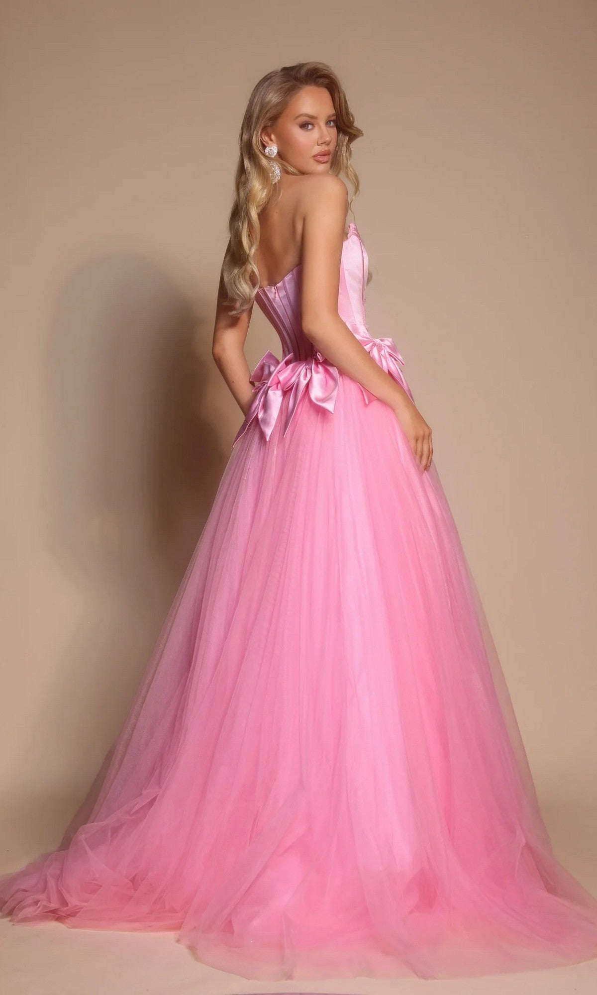 A woman wearing the Portia and Scarlett PS26102 strapless ball gown with a corset bodice and tulle skirt stands against a beige background, facing slightly away from the camera.